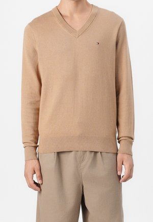 Mężczyzna ubrany w beżowy sweter z dekoltem w serek z małym logo Tommy Hilfiger na piersi, w połączeniu z beżowymi spodniami, ręce swobodnie opuszczone wzdłuż ciała.