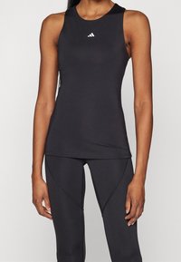 Kvinna iklädd en ärmlös svart Adidas träningslinne och matchande svarta leggings med synliga sömmar, mot en enkel bakgrund.