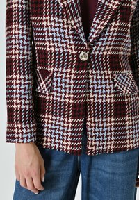 Blazer en tissu texturé arborant un motif à carreaux rouge, bleu et crème, avec fermeture à un bouton et deux poches avant.