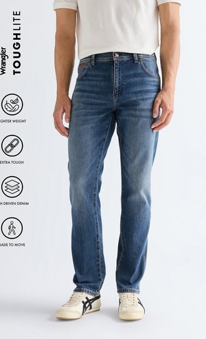 Hombre con jeans azules de corte recto y zapatillas crema, de pie contra un fondo claro y sencillo con la marca Wrangler Toughlite y texto de características.