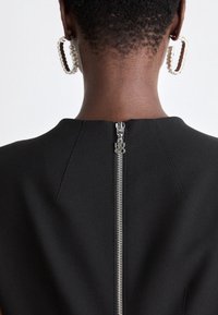 Gros plan sur le cou et le dos d'une personne portant un vêtement noir avec une fermeture éclair centrale argentée et des boucles d'oreilles rectangulaires distinctives.