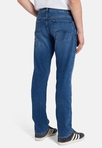 Jeans in denim blu con design a gamba dritta, caratterizzati da un lavaggio medio, dettagli di cucitura sulle tasche posteriori e indossati con sneakers bianche e nere.