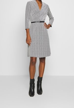 Femme portant une robe longueur genou à motif chevron noir et blanc, manches trois-quarts, ceinture noire et bottines noires.