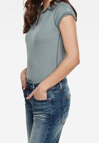Camisa polo azul claro de manga corta, con cuello y un diseño ajustado. Combinada con unos jeans de denim de lavado medio, que cuentan con bolsillos frontales y detalles desgastados.