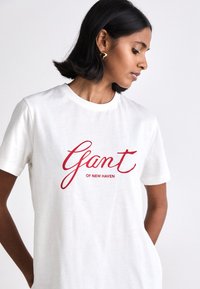 Biała bawełniana koszulka z krótkim rękawem, z okrągłym dekoltem, z luźnym krojem, z czerwonym kursywnym napisem "Gant" nad napisem "OF NEW HAVEN."
