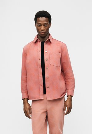 Man draagt een koraalroze overhemd met bloemenpatroon over een zwart T-shirt en bijpassende koraalroze broek, staand tegen een effen witte achtergrond.