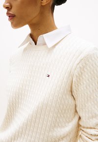 Ženska, ki nosi belo pulover z vzorcem kable, na prsih majhen rdeče-bel-modri logotip, pod njim pa bela srajca z ovratnikom.