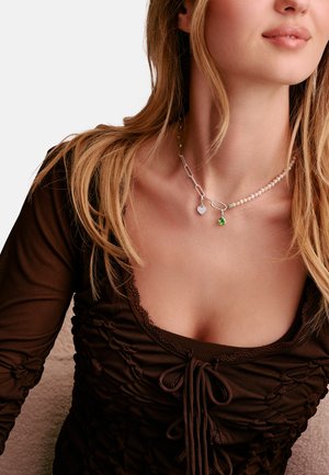 Junge Frau mit langen blonden Haaren trägt ein braunes, gerafftes Oberteil und eine Halskette aus gemischten Materialien mit Herz- und grünem Edelsteinanhänger.