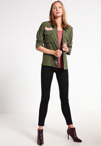 Olivengrønn militærjakke med lapper, sammen med en burgunder topp, svarte skinny jeans og burgunder ankelstøvler. Glatt stoff, tettsittende design.