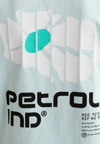 Jasnoniebieska koszulka z dużym białym abstrakcyjnym wzorem, turkusowym akcentem i wyrazistym czarnym napisem "petrol ind" oraz szczegółami dotyczącymi marki.