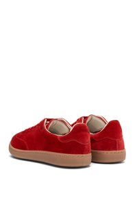 Czerwone zamszowe trampki mają okrągły nosek, sznurowany design, gumową podeszwę oraz kontrastujące beżowe akcenty na podeszwie i kołnierzu.