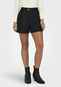 Schwarze strukturierten Shorts mit hoher Taille, vorderem Knopfverschluss und taillierter Passform. Kombiniert mit schwarzen Knöchelstiefeln und einem cremefarbenen Langarmoberteil.