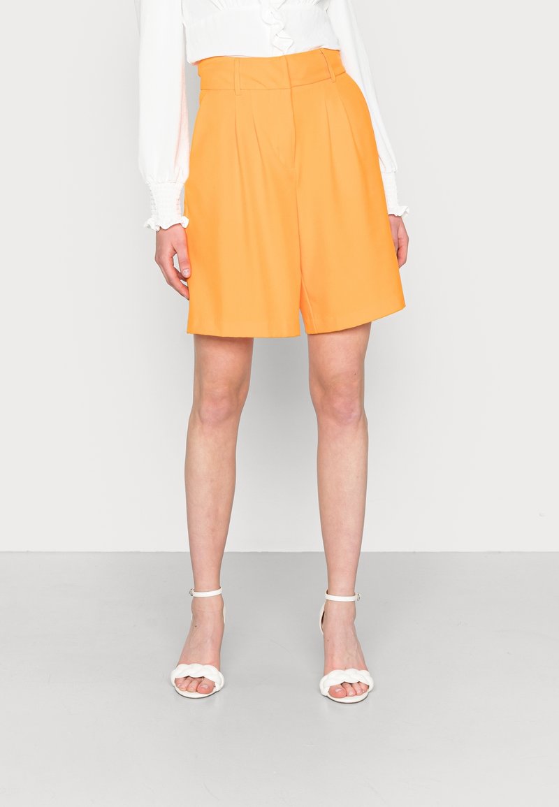 Femme portant un short taille haute, orange vif, longueur genou avec des plis, associé à un chemisier blanc et des sandales blanches à bride chevillère sur un sol gris.