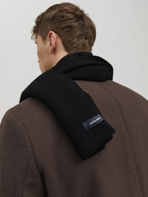 Black knitted scarf draped across a brown coat, featuring a textured finish and a small brand label on the scarf's edge.