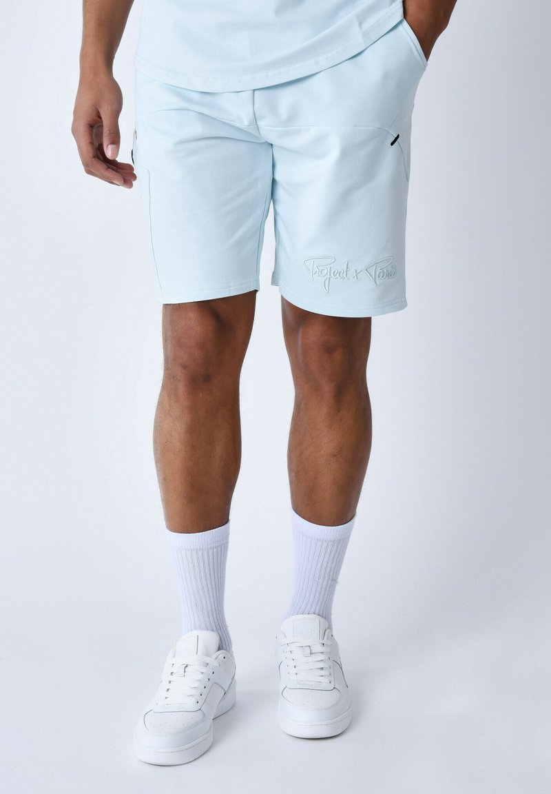 Pantaloni corti azzurri realizzati in un tessuto morbido, con tasche laterali e un logo decorativo ricamato di un colore a contrasto. Abbinati a sneakers bianche e calze.