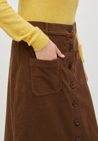 Jupe en coton brun avec un devant à boutons, des poches latérales et une texture douce. Le modèle porte un pull jaune à manches longues.
