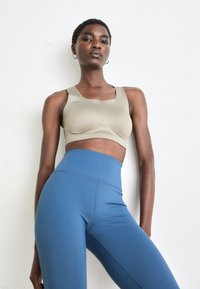 Selvsikker kvinne i beige sports-bh og høyhalsede blå tights poserer mot en enkel hvit vegg, med hendene avslappet ved sidene.