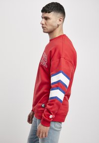 Roter Kapuzenpullover mit gerippten Bündchen, der weiße und blaue Streifenakzente an den Ärmeln und ein Logo auf der Brust trägt. Weicher Stoff.