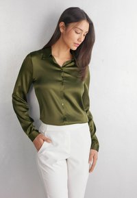 Blusa di raso verde oliva con colletto, chiusura con bottoni e maniche lunghe, abbinata a pantaloni bianchi su misura. È visibile una consistenza liscia.