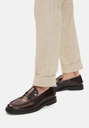Brązowe skórzane loafersy z błyszczącym wykończeniem, ozdobione przeszyciami i dekoracyjnymi detalami, zestawione z beżowymi spodniami o fakturowanej tkaninie.