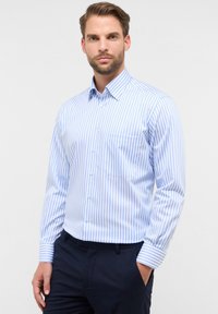 Uomo con barba che indossa una camicia a righe verticali blu e bianche e pantaloni scuri, in piedi con una mano in tasca, sfondo neutro.