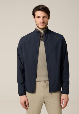 Jeune homme portant une veste zippée marine par-dessus un pull en maille beige et un pantalon kaki clair, debout devant un fond neutre uni.