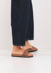 Sandales à brides à motifs marron avec une semelle intérieure en liège, ornées d'une boucle décorative, portées avec un pantalon en jean foncé raccourci.