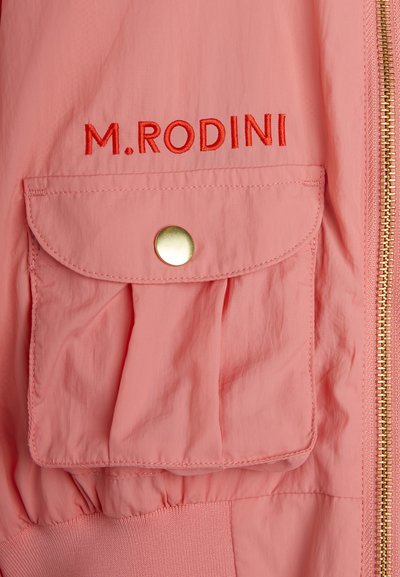 Gaiši rozā jaka ar zelta rāvējslēdzēju, priekšējo kabatu ar spiedpogu un izšūtu "M. RODINI" sarkaniem burtiem. Izgatavota no gluda auduma.