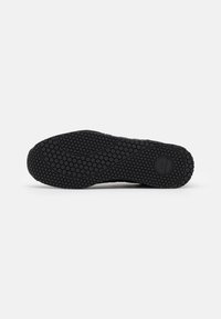 Semelle de chaussure en caoutchouc noir avec motif de bande de roulement hexagonal, rainures courbes et détail de logo circulaire sur fond blanc lisse.