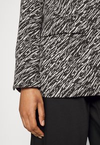 Zbliżenie na dłoń obok kieszeni marynarki w czarno-białe wzory oraz czarnych spodni.