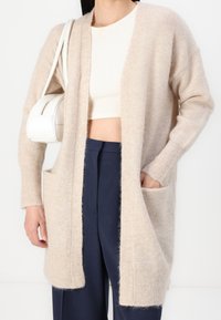 Béžový pletený kardigan s otevřeným předním dílem, dlouhými rukávy a bočními kapsami. Měkká textura a volný střih v kombinaci s tmavě modrými kalhotami.