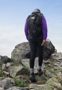 Sac à dos de randonnée noir avec des sangles réglables, logo à l'avant et design en cordon bungee. Personne portant un pull en polaire violet et des leggings noirs, marchant sur un terrain rocheux.