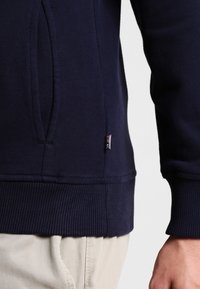 Sudadera azul marino con una textura suave, dobladillo acanalado y bolsillo lateral. Etiqueta de logo pequeña en la costura inferior, que contrasta con el material.
