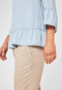 Top azzurro chiaro a maniche lunghe con orlo arricciato e texture morbida. Abbinato a pantaloni beige, mano visibile di lato.