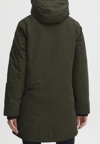 Armádní zelená parka do délky kolen s kapucí, s hladkým povrchem a elastickými manžetami. Obsahuje postranní logo jako akcent. Zobrazeno zádové vyobrazení.