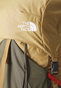 Olíva zöld és bézs szövet hátizsák textúrázott felülettel, a The North Face logójával és állítható pántokkal, különböző anyagokat bemutatva.