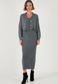 Vestido midi de punto gris con textura de canalé, que incluye un cárdigan corto con tres botones dorados. Combinado con botas negras hasta el tobillo.