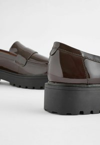 Mocassins en cuir brun foncé brillant avec semelles épaisses en caoutchouc noir à crampons, vus de biais et de l'arrière en contre-plongée.