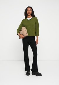 Green ribbed cardigan with buttons, worn over a white shirt. Paired with black flared pants and black platform shoes. Holding a beige bag.