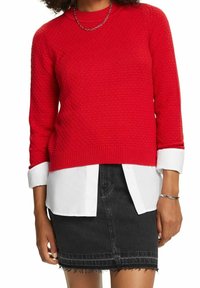 Pull rouge en maille avec un motif texturé, doté d'un col et de poignets côtelés, porté sur une chemise blanche à col, associé à une jupe en denim noire.