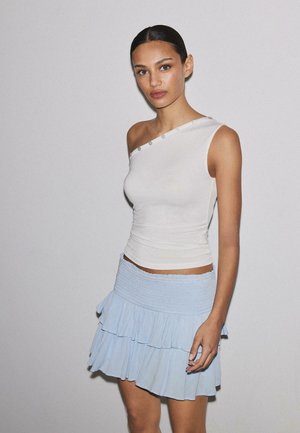 Kobieta z gładkimi ciemnymi włosami ubrana w biały top na jedno ramię z guzikami oraz jasnoniebieską, kilkuwarstwową mini spódnicę, stoi na tle gładkiej jasnej ściany.