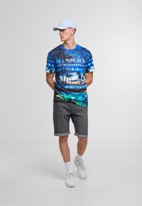 Blue graphic t-shirt with palm tree and sunrise design, paired with gray denim shorts. Model wears white sneakers and a light blue cap.