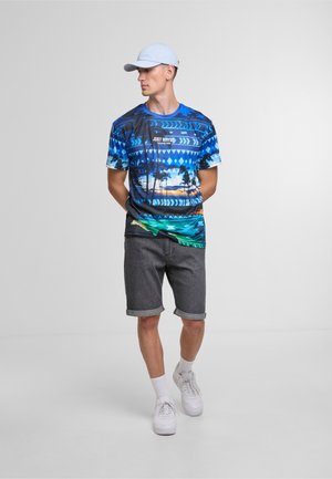 Blue graphic t-shirt with palm tree and sunrise design, paired with gray denim shorts. Model wears white sneakers and a light blue cap.