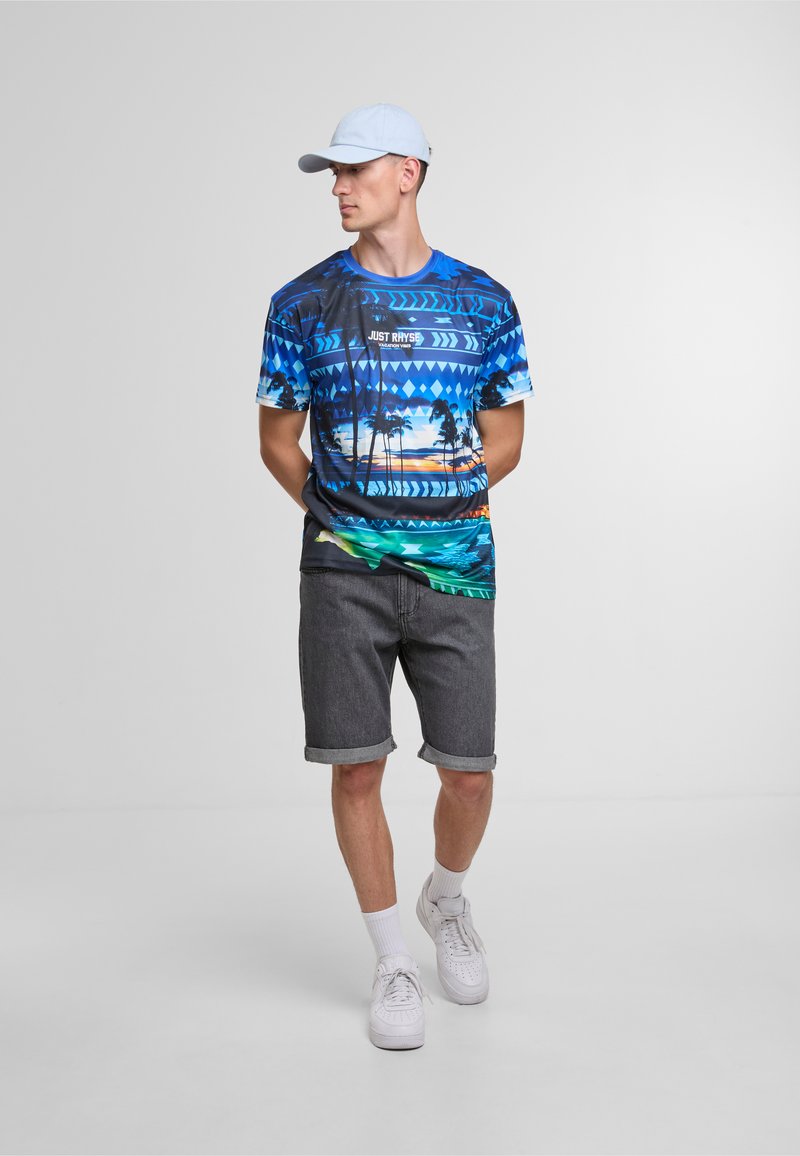 Blå grafisk t-skjorte med palme- og soloppgangsdesign, kombineres med grå denimshorts. Modellen har på seg hvite joggesko og en lys blå caps.