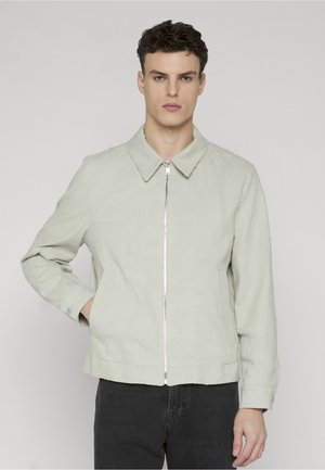 Joven con chaqueta gris claro con cremallera y cuello, de pie con una mano en el bolsillo, frente a un fondo claro y uniforme.