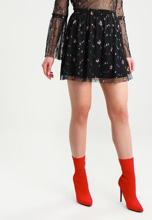 Mini-jupe noire en tissu transparent avec broderie florale, associée à des bottines rouges vives avec un bout pointu et un talon haut.