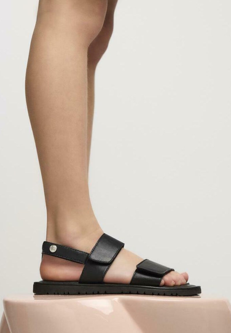 Sandalias de cuero negro con correas ajustables, suelas planas y un diseño minimalista. Integración sin costuras de acentos metálicos en las correas.