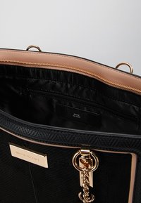 Musta River Island -käsilaukku, jossa on kultaketju, beige reunus ja sisäinen vetoketjupussi tasaisella harmaalla taustalla.