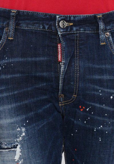 Jeans de denim azul oscuro con secciones desgastadas, salpicaduras de pintura blanca y roja, costuras visibles y una etiqueta de marca en la cintura.