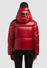 Sarkana pūkaina jaka ar spīdīgu, kvadrātveida faktūras ārpusi, kapuci un logotipu aizmugurē. Raksturo plašas piedurknes un īsu siluetu.